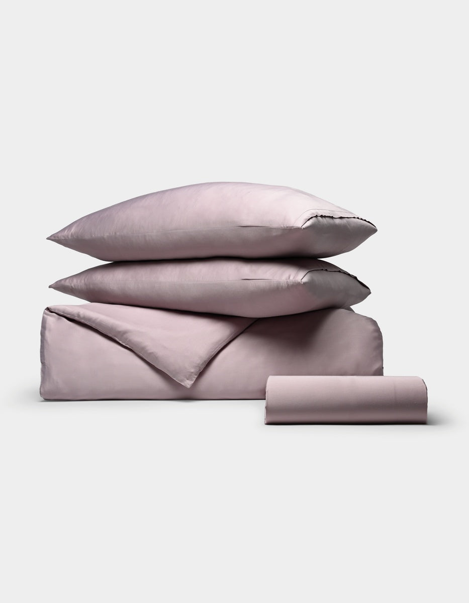 The Cozy Earth Bamboo Bedding Essential Bundle is displayed on a plain white background, featuring a neatly stacked set of light mauve bedding that includes two pillows, a folded sheet, and a rolled sheet.