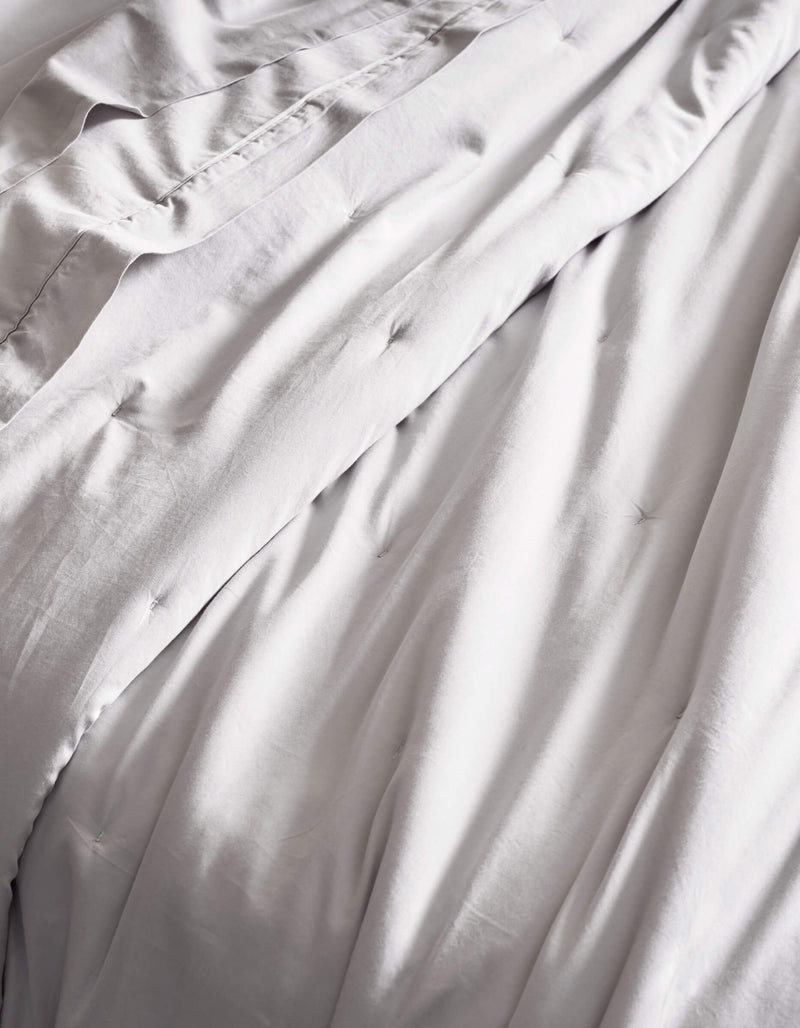 The Cozy Earth Soft-Wash Cotton Quilt features smooth, light gray bedding with gentle folds and subtle tufted details, offering a soft, inviting texture.