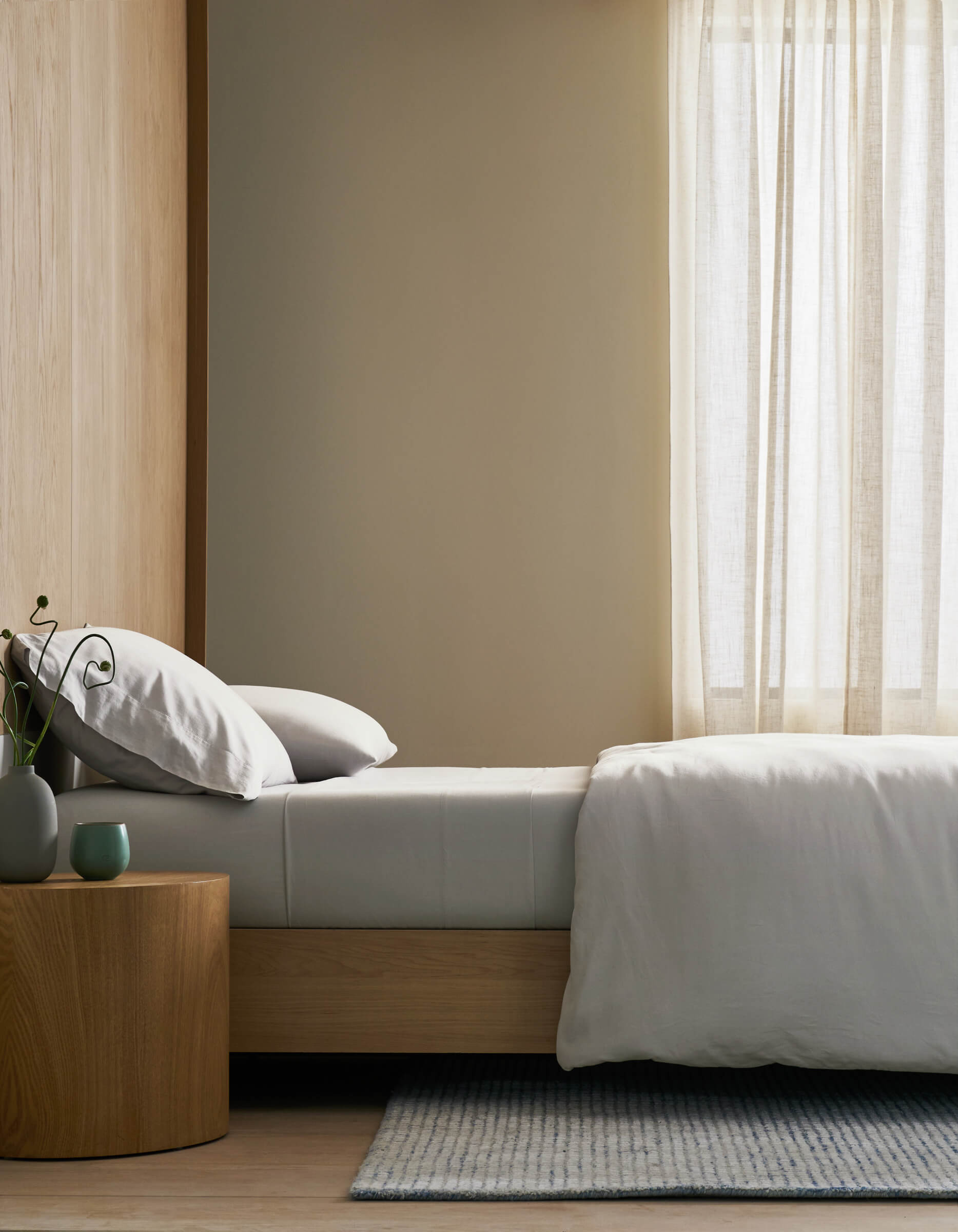 Minimalist bedroom featuring a neatly made bed with Cozy Earth’s Soft-Wash Cotton Duvet Cover, white bedding, a wooden nightstand adorned with a small plant and vase, light walls, and sheer curtains letting in natural light.