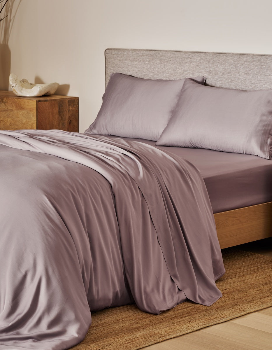 A bed dressed in a silky mauve-lavender bedding set with matching pillowcases, styled in a warm bedroom setting with a wooden nightstand and textured headboard. 