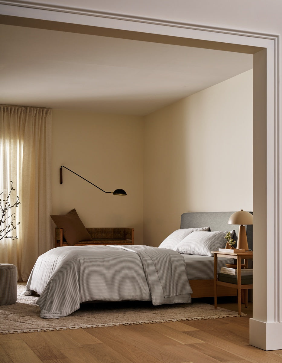A minimalist bedroom interior with a neatly made bed dressed in white linens, a gray upholstered headboard, warm beige walls, wood flooring, a bedside table with a lamp, and a wall-mounted black lamp. 
