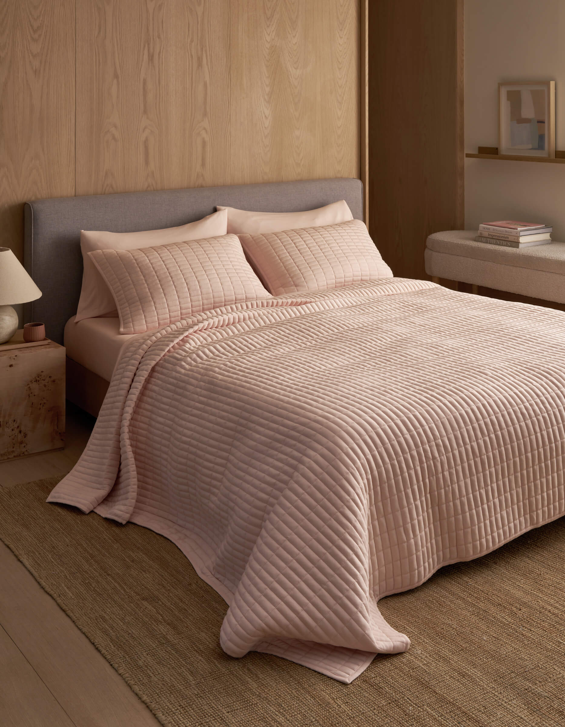 A neatly made bed features the Cozy Earth Bamboo Jersey Quilt in light pink with matching pillows, set against a wooden wall in a minimal, modern bedroom with neutral tones and simple decor.