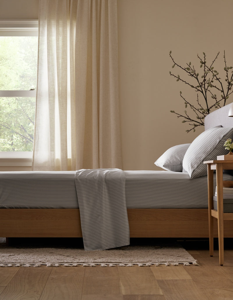 A bed neatly made with the Cozy Earth Bamboo Flat Sheet in gray stripes sits beside a wooden nightstand, set in a softly lit room with light curtains, a window, and a decorative plant branch in the background.