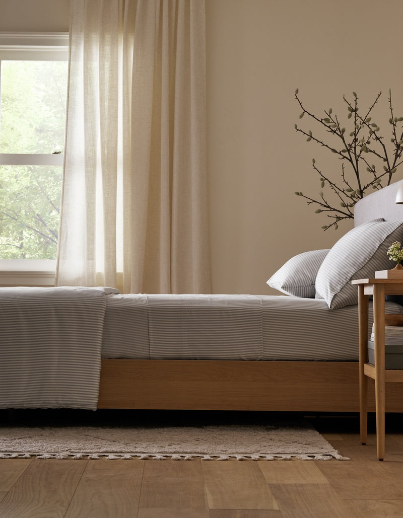 A neatly made bed featuring the Cozy Earth Bamboo Fitted Sheet sits beside a wooden nightstand, a branch in a vase, and a window with sheer curtains letting in natural light. Light wood floors and a rug complete the cozy scene.