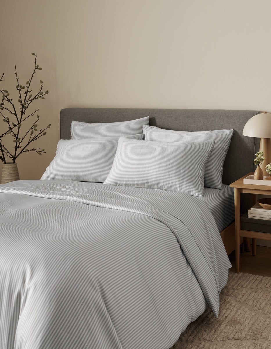 A neatly made bed features the Bamboo Bedding Ultimate Bundle by Cozy Earth, showcasing light gray striped linens and a gray upholstered headboard topped with two pillows. Beside the bed, a wooden nightstand holds a lamp, books, and a vase, while a decorative branch accents the corner against a beige wall.