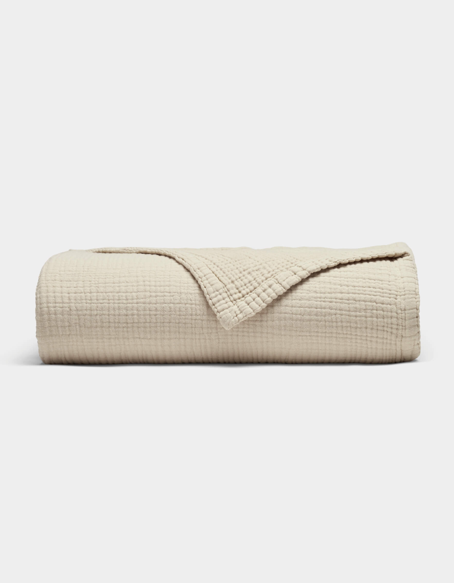 The Cozy Earth Cotton Gauze Blanket, in beige with a soft grid texture, is neatly rolled with a corner folded back to reveal its fabric, shown against a plain white background.|Color:Coconut