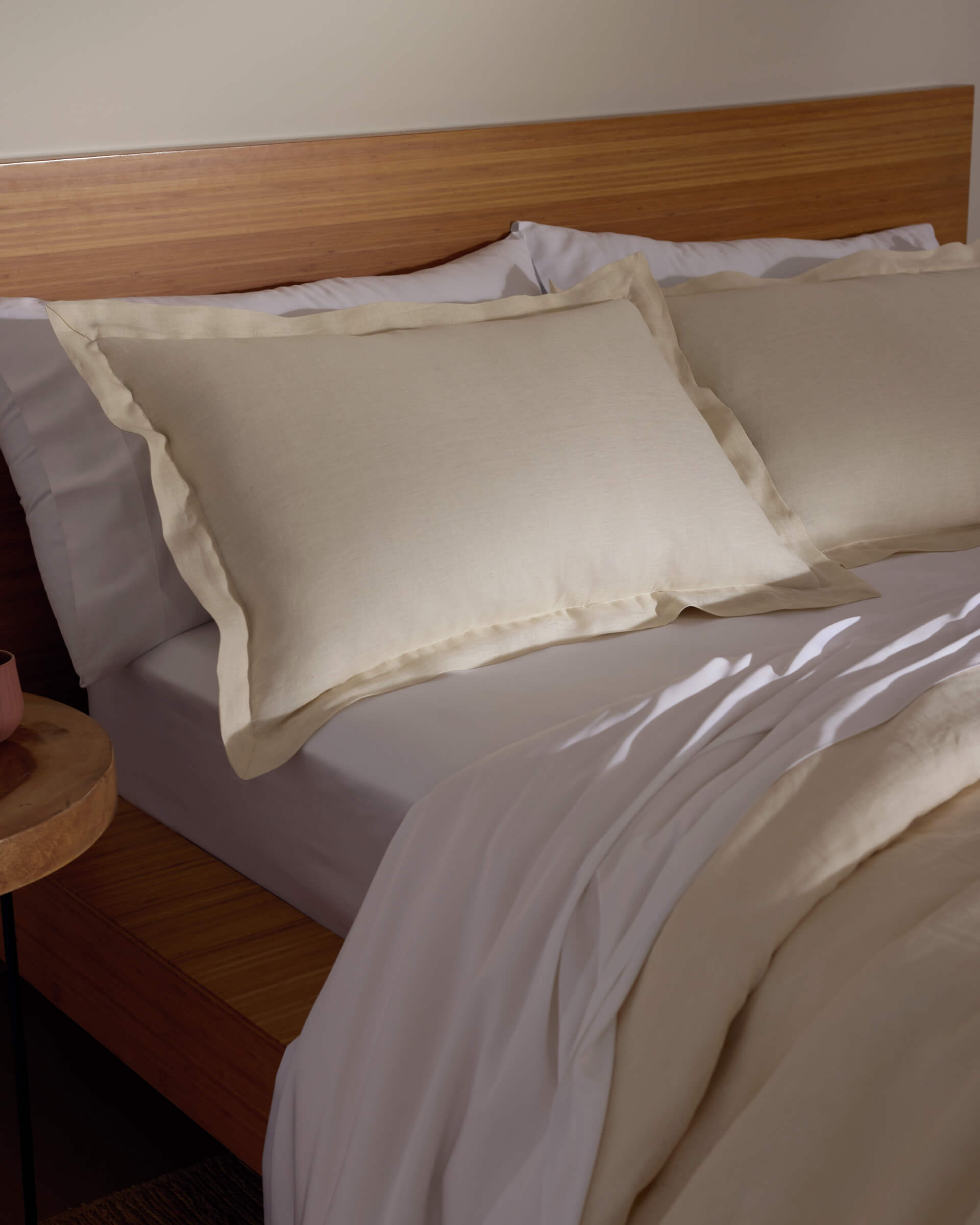 A bed featuring Cozy Earth's Linen Shams in cream, paired with a matching comforter, sits against a wooden headboard and nightstand, all bathed in soft natural light.|Color:Flax