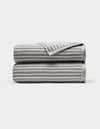 Two neatly folded Ribbed Terry Bath Towels by Cozy Earth, light gray with horizontal white stripes, stacked atop each other against a plain white background. A corner of the top towel is turned over, revealing the underside.|Color:Dew Stripe
