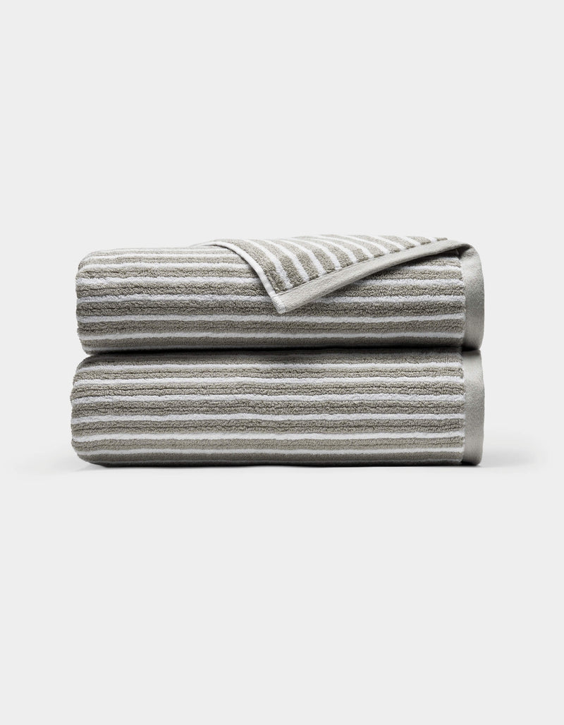 Two neatly folded Ribbed Terry Bath Towels by Cozy Earth, light gray with horizontal white stripes, stacked atop each other against a plain white background. A corner of the top towel is turned over, revealing the underside.