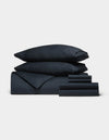 The Cozy Earth Soft-Wash Cotton Bedding Core Bundle, featuring two stacked pillows, a duvet, and several sheets in dark navy blue, is neatly arranged against a plain white background.|Color:Eclipse