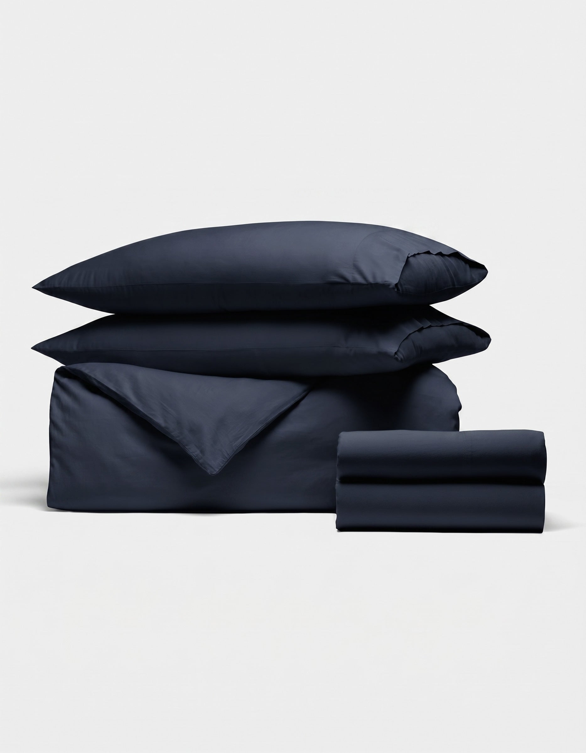 The Soft-Wash Cotton Bedding Deluxe Bundle by Cozy Earth features two pillows, a folded duvet, and sheet set in dark navy blue, neatly stacked against a plain white background.|Color:Eclipse