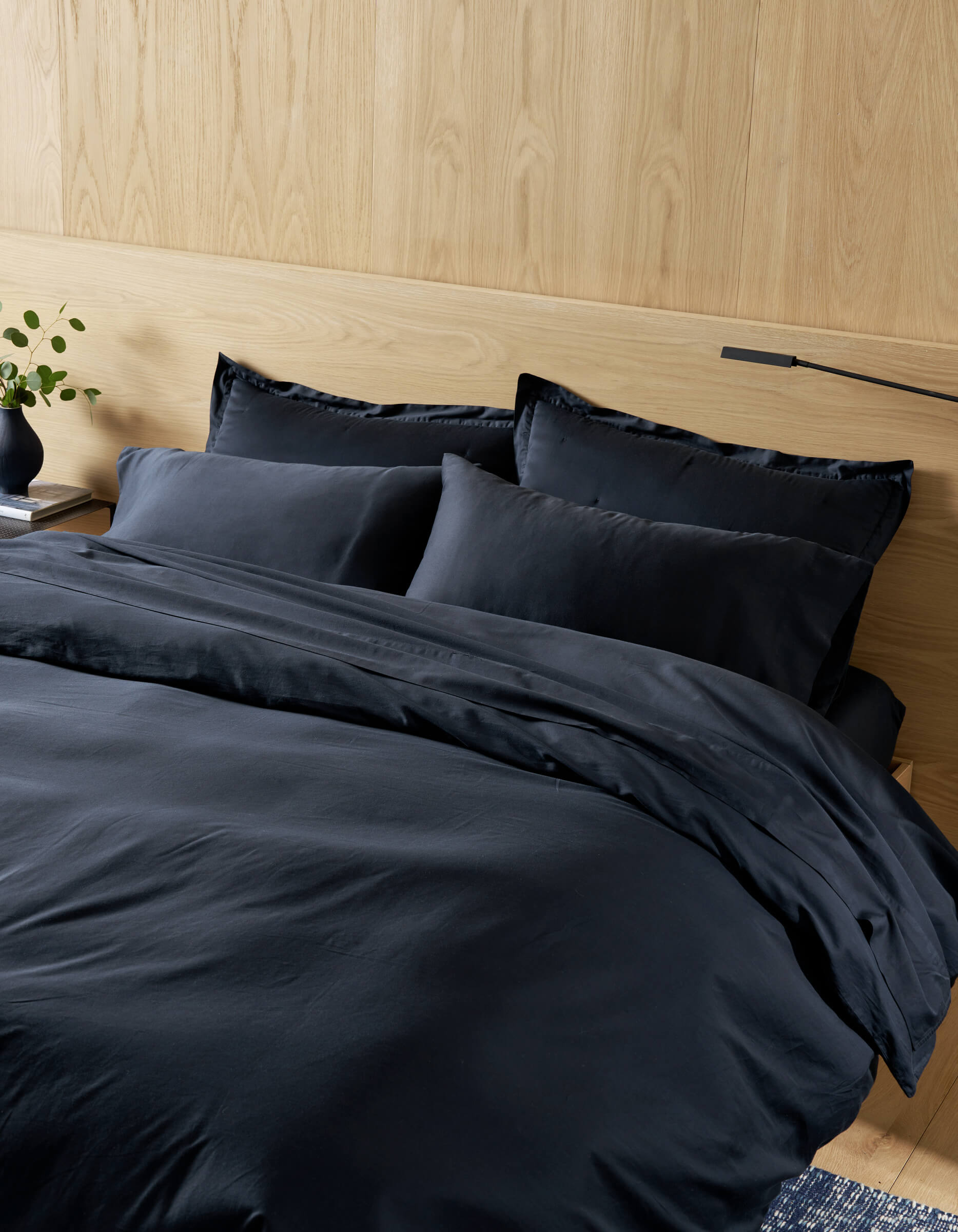 A neatly made bed features Cozy Earth Soft-Wash Cotton Pillowcases in dark navy, along with matching bedding and a comforter, against a light wooden headboard. A vase with green stems rests on the bedside table.