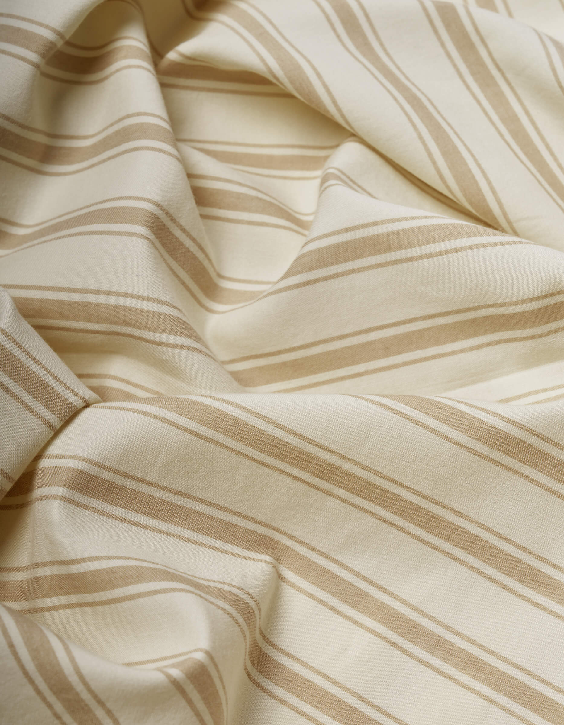 The Cozy Earth Soft-Wash Cotton Flat Sheet features cream-colored fabric with light brown diagonal stripes, softly draped with gentle folds and subtle shadows across its slightly wrinkled surface.|Color:Coconut and Sandalwood Stripe