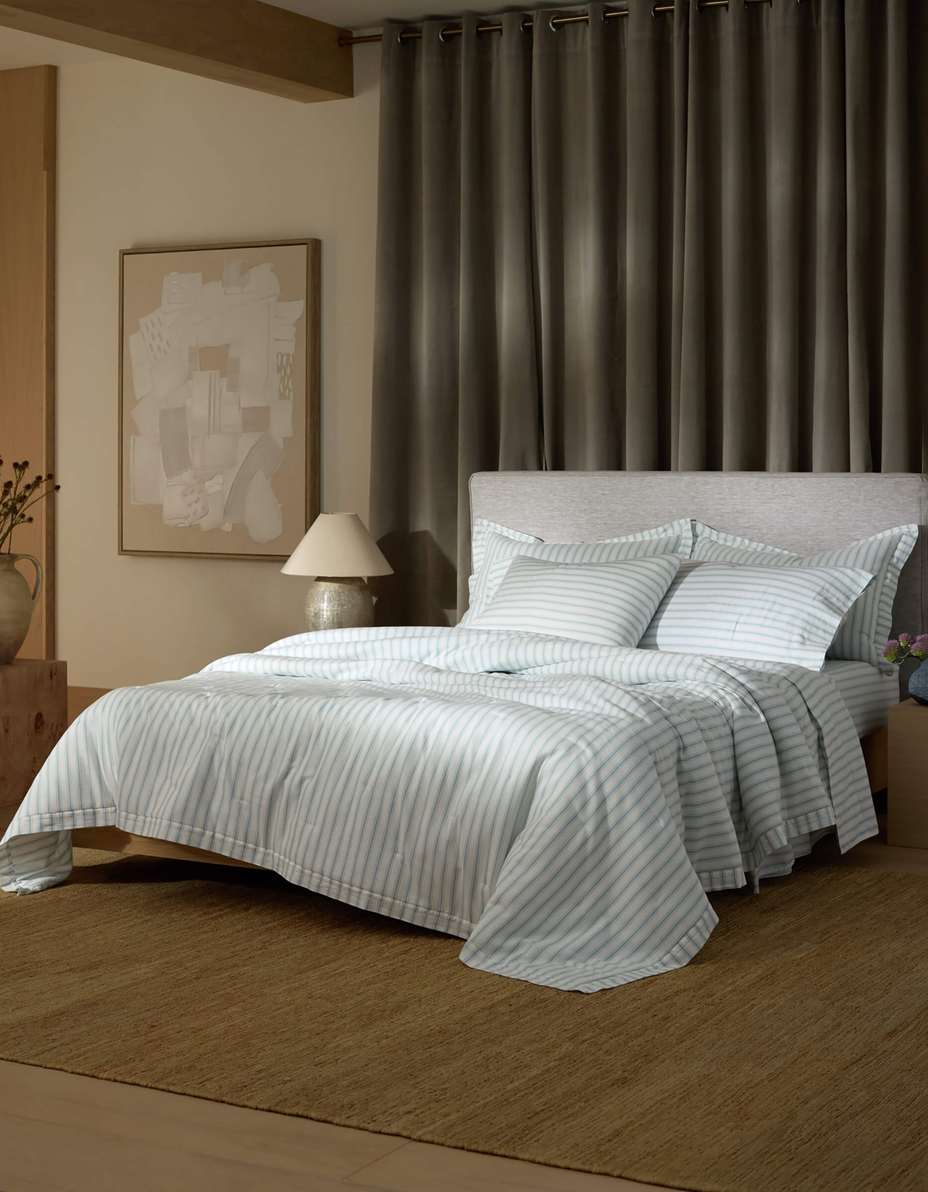 A modern bedroom features the Cozy Earth Soft-Wash Cotton Quilt in light blue and white stripes, set on a neatly made bed with a gray upholstered headboard, abstract wall art, bedside table with lamp and vase, gray curtains, and a woven rug.