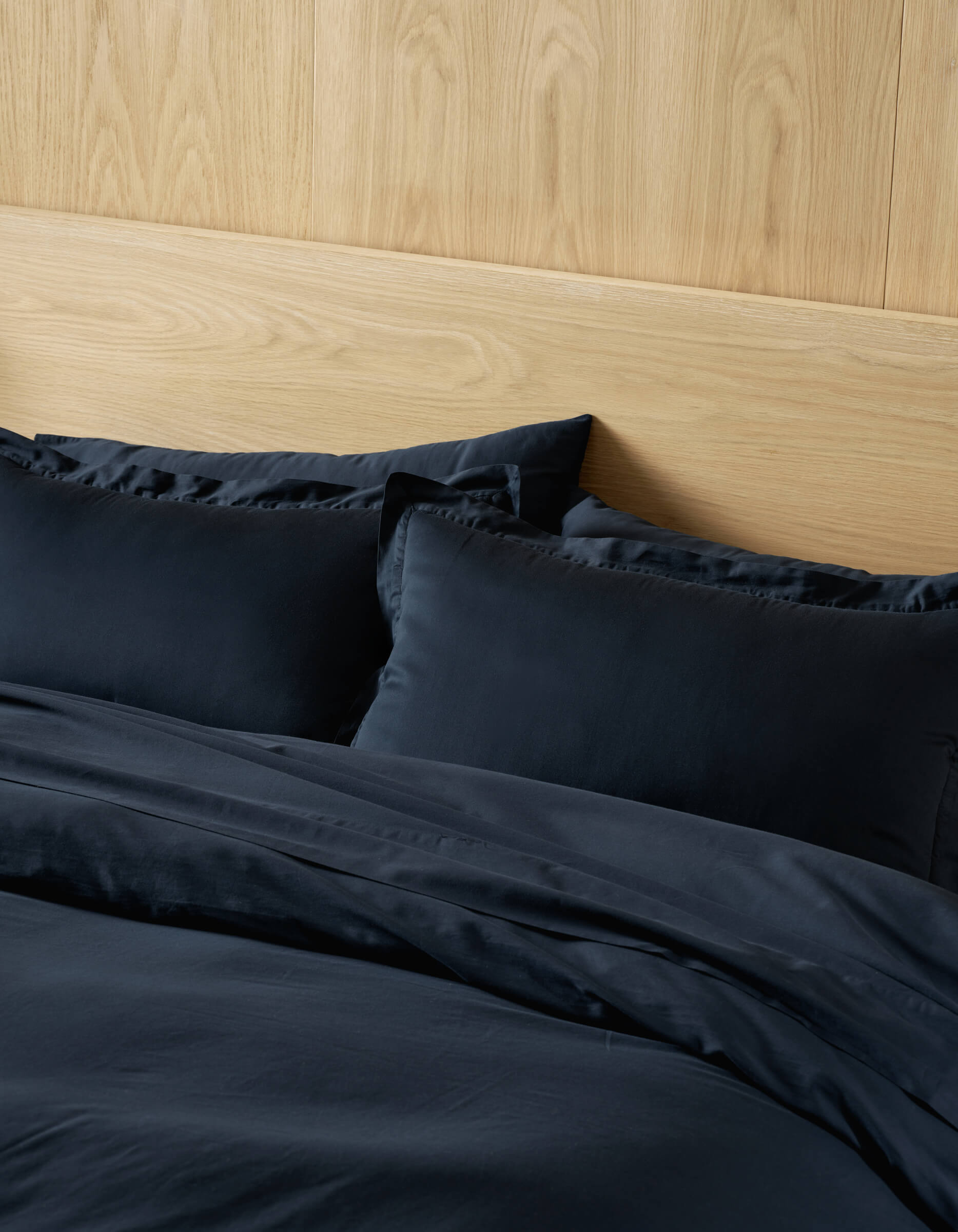 A neatly made bed with Cozy Earth Soft-Wash Cotton Shams in dark navy, paired with matching sheets, stands against a light wooden headboard and a wood-paneled wall.
