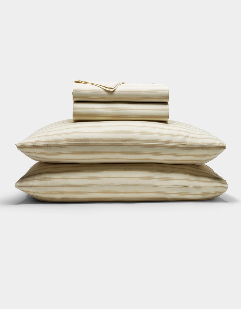 The Cozy Earth Soft-Wash Cotton Sheet Set in beige and light brown stripes is neatly stacked with two pillows at the base, topped with folded sheets and a pillowcase, displayed against a plain white background.