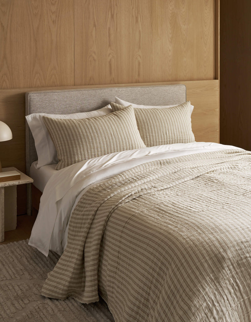 The cozy, minimalist bedroom features a Waterfront Coverlet Bundle by Cozy Earth on the bed with two pillows, set against a light wood panel wall. A small bedside table with a white lamp and a textured rug complete the scene.