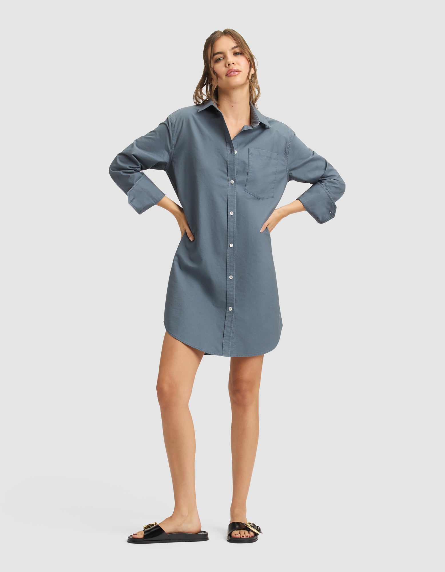 A woman wears the Cozy Earth Women's Camden Dress, a loose-fitting blue button-down shirt-dress, paired with black slide sandals with gold accents. Her long hair is pulled back and she stands against a plain light background. 