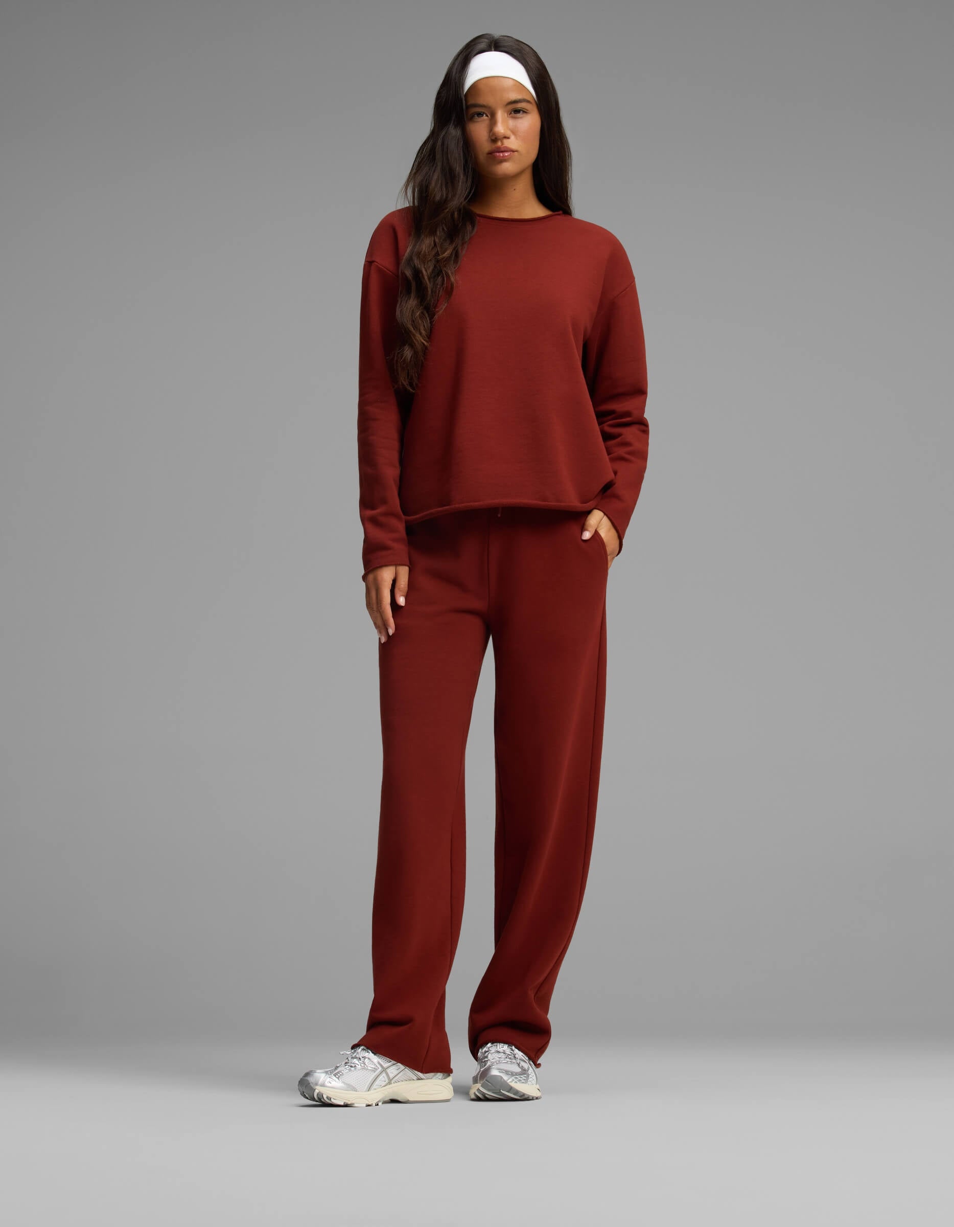 A woman stands against a plain gray background wearing the Cozy Earth Women's Chelsea Set—a matching rust-colored long-sleeve top and pants. She has long dark hair, a white headband, and white sneakers, with one hand in her pocket.