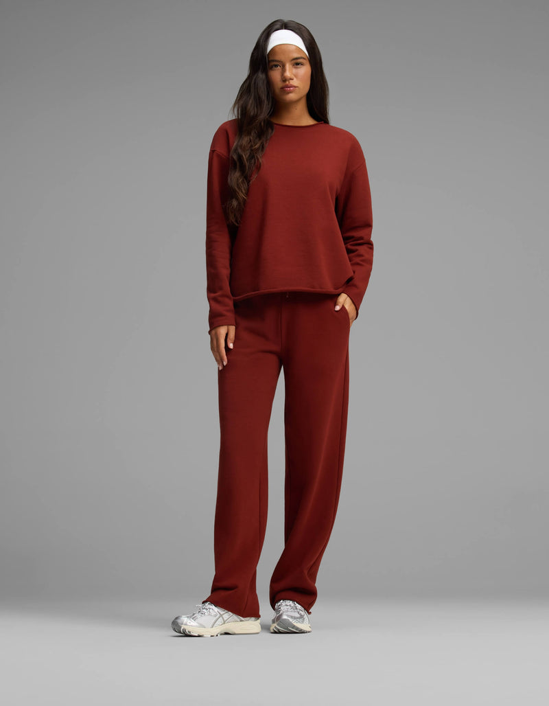 A woman stands against a plain gray background wearing the Cozy Earth Women's Chelsea Set—a matching rust-colored long-sleeve top and pants. She has long dark hair, a white headband, and white sneakers, with one hand in her pocket.