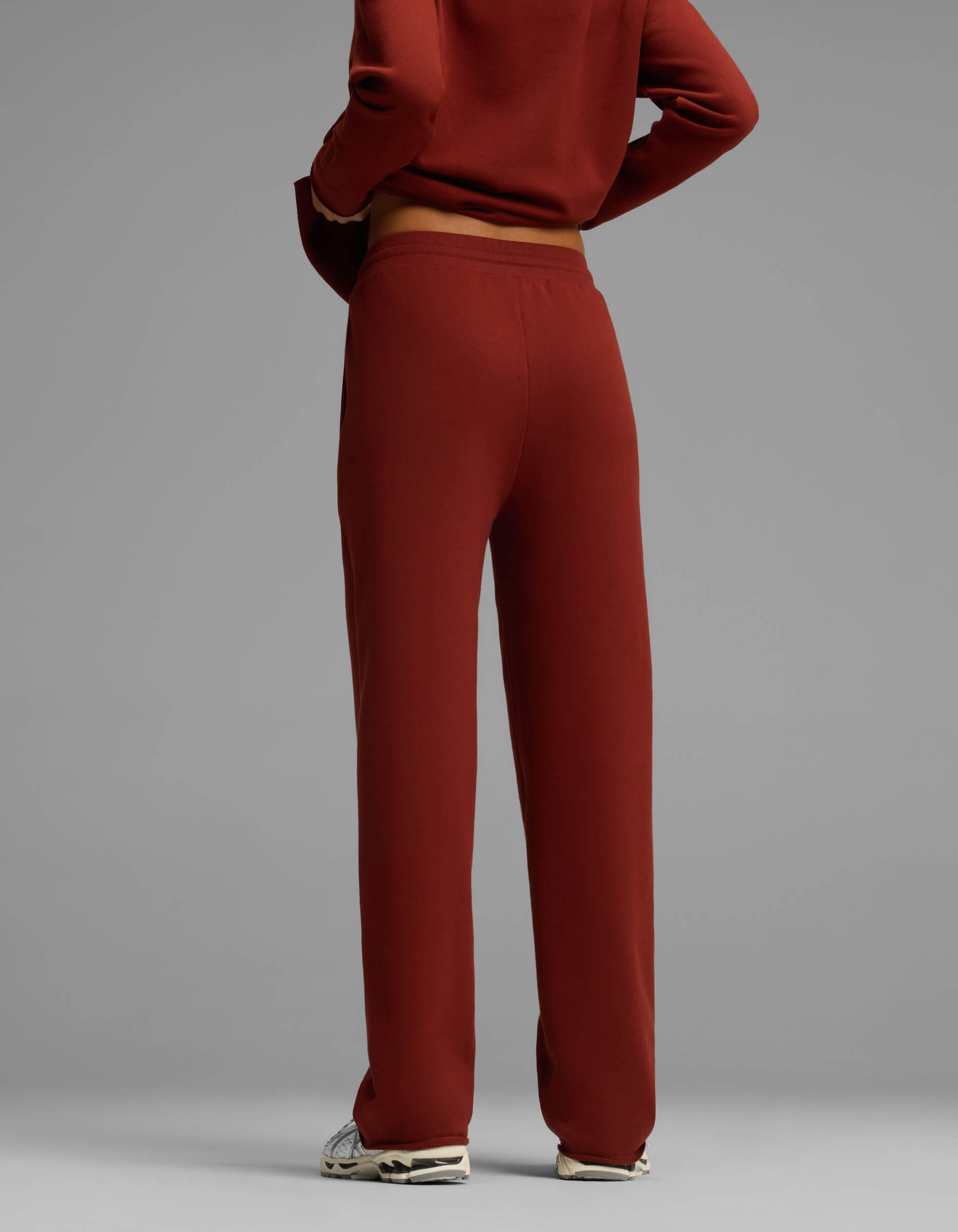 A woman is shown from the back wearing Cozy Earth's Women's Chelsea Pant in a red-brown hue, paired with a matching top. The image focuses on her lower torso and legs, with arms crossed, set against a plain gray background.|Color:Currant
