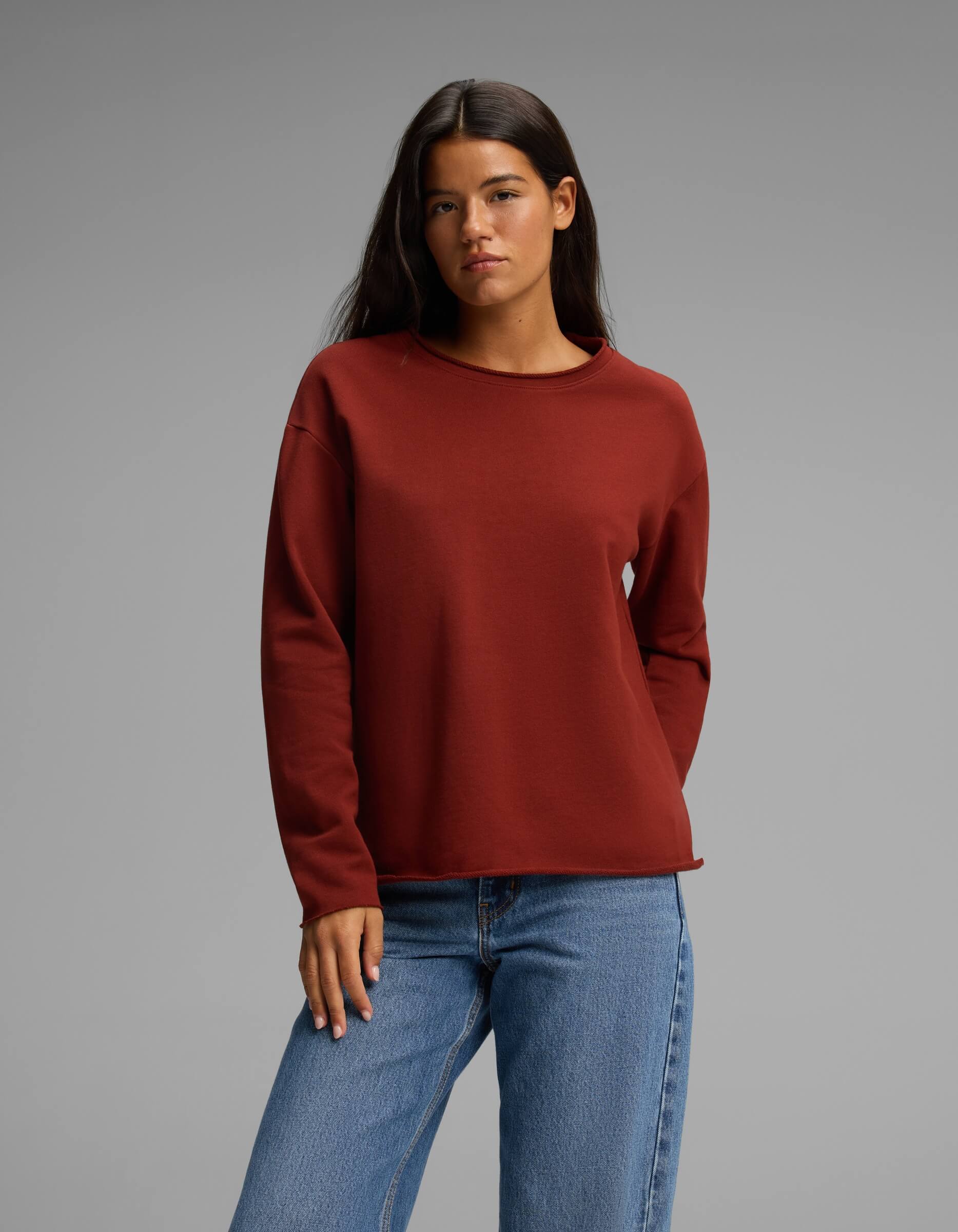 A woman with long dark hair wears the Cozy Earth Women's Chelsea Pullover in a rust color paired with blue jeans, standing against a plain gray background with a neutral expression.|Color:Currant