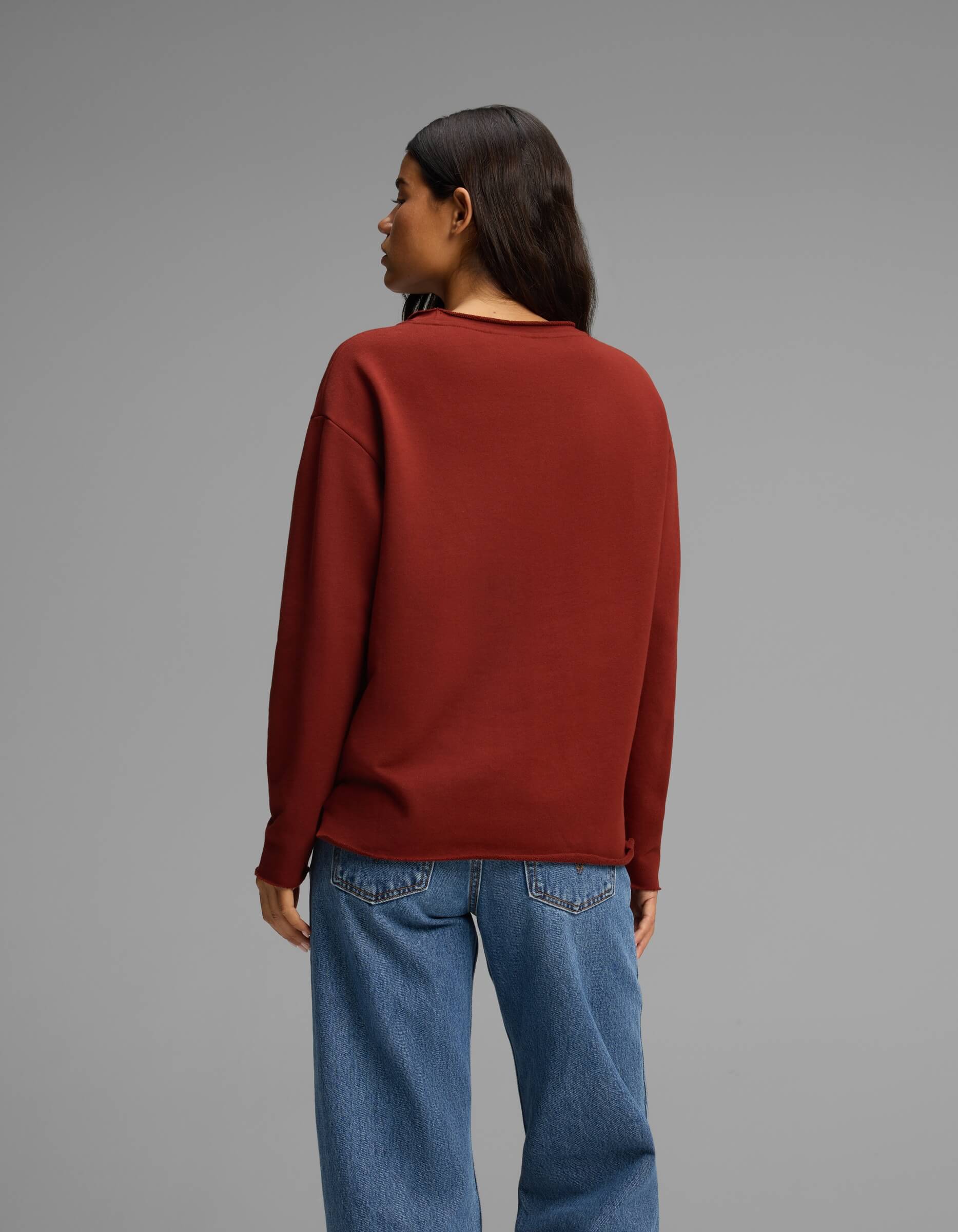 A woman with long dark hair, wearing the Cozy Earth Women's Chelsea Pullover in maroon and blue jeans, stands facing away from the camera against a plain gray background.|Color:Currant