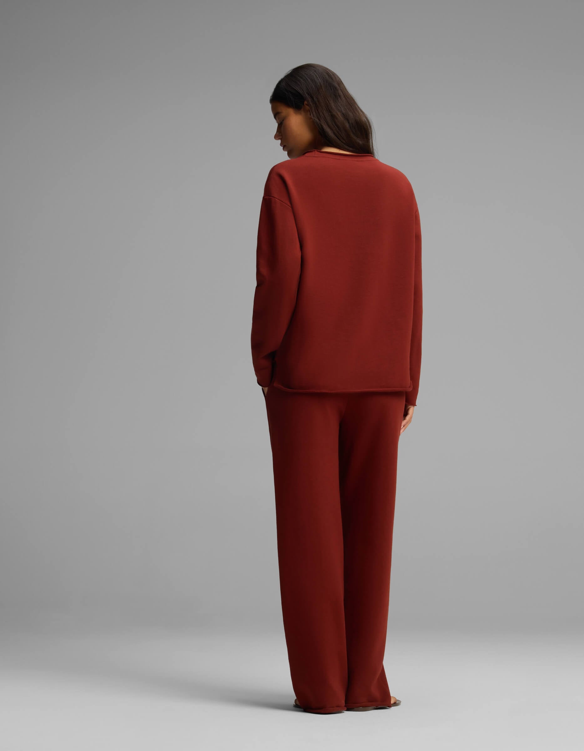 A person with long dark hair stands facing away, wearing the Cozy Earth Women's Chelsea Set—a loose-fitting, long-sleeve dark red top and matching wide-leg pants—against a plain gray background.