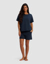 A woman with long dark hair stands barefoot against a plain light background, wearing the Cozy Earth Women's Chelsea Short Sleeve Set in dark navy, with her hands in her pockets.|Color:Eclipse