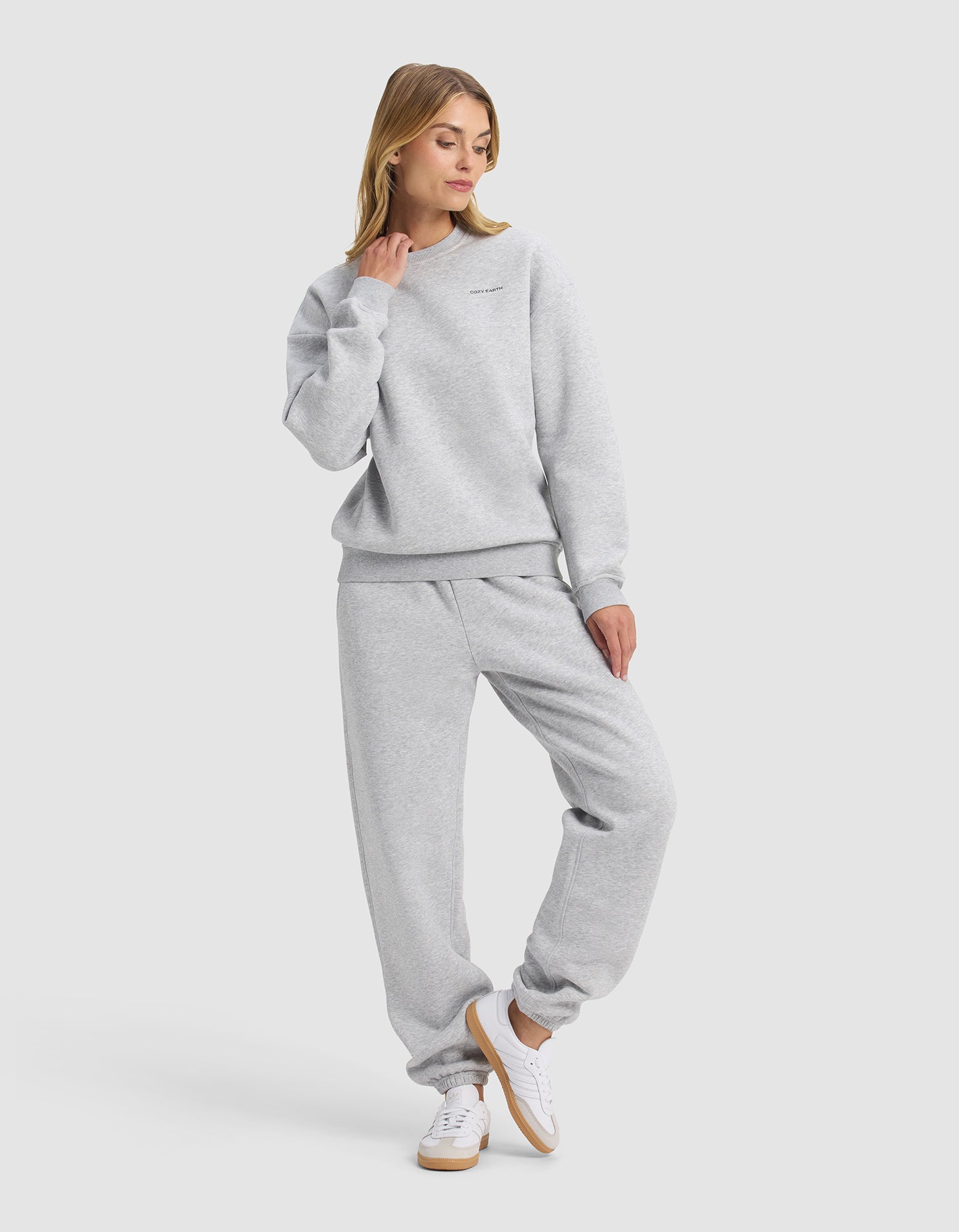 A woman models Cozy Earth’s Women's Cityscape 2.0 Crewneck & Sweatpant Set in light gray, paired with white sneakers, standing casually with one hand on her neck and gazing slightly to the side.|Color:Heather Grey