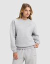 A woman with long blonde hair models the Cozy Earth Women's Cityscape 2.0 Crewneck in light gray, paired with matching sweatpants, standing with one hand in her pocket against a plain light background.|Color:Heather Grey