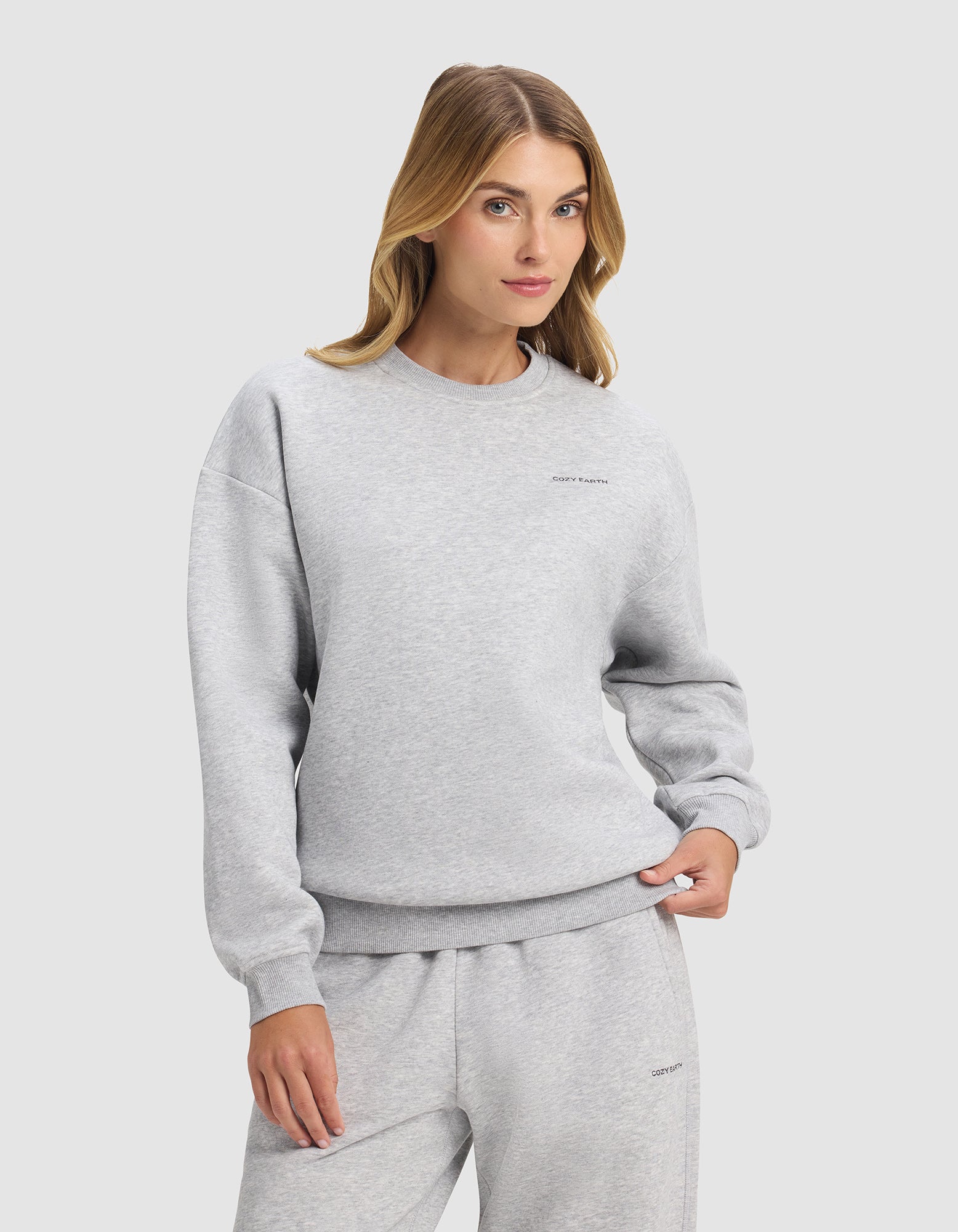 A woman with long blonde hair models the Cozy Earth Women's Cityscape 2.0 Crewneck in light gray, paired with matching sweatpants, standing with one hand in her pocket against a plain light background.|Color:Heather Grey