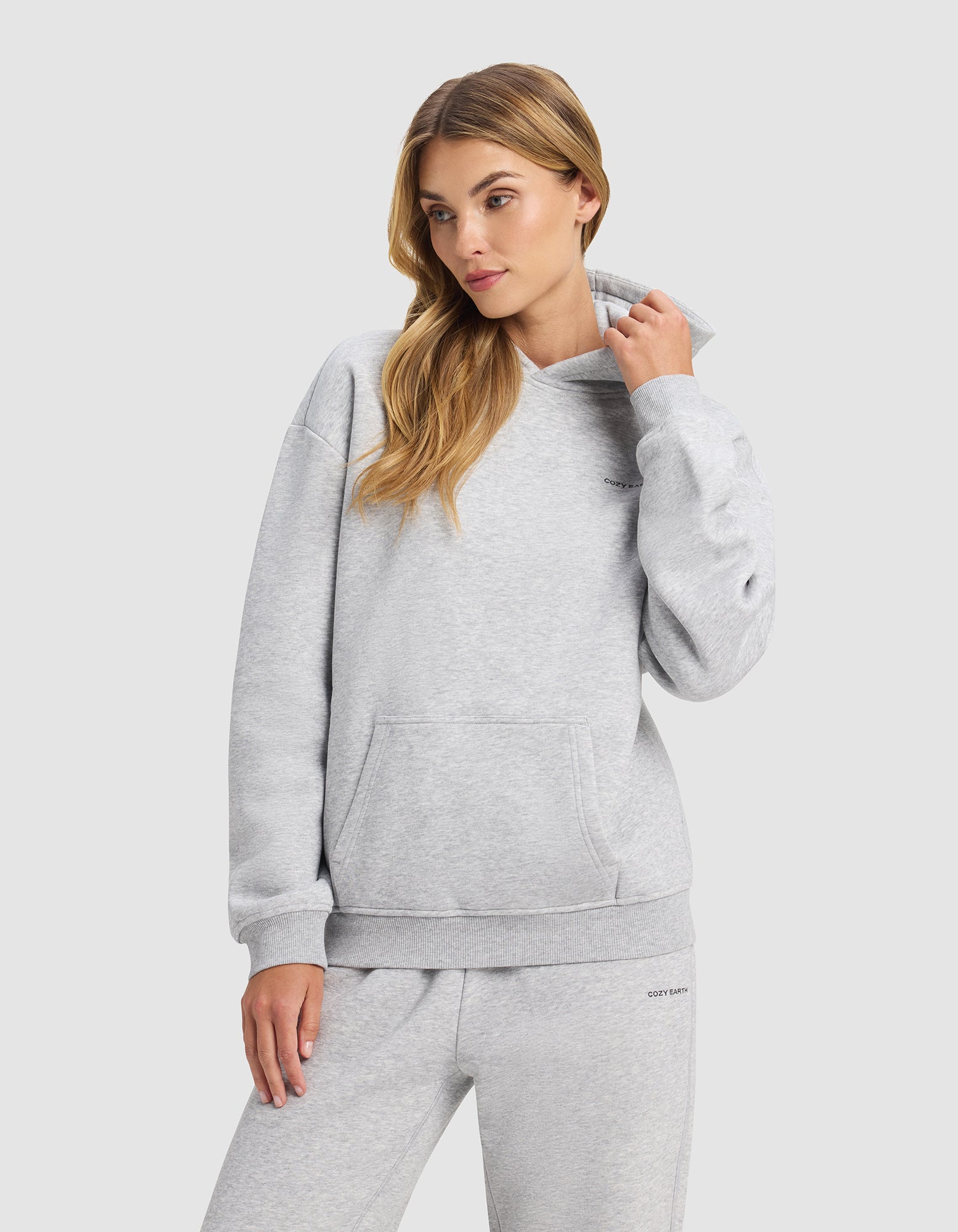 A woman wears the Cozy Earth Women's Cityscape 2.0 Hoodie in light gray, standing against a plain background and looking to the side as she adjusts her collar.|Color:Heather Grey