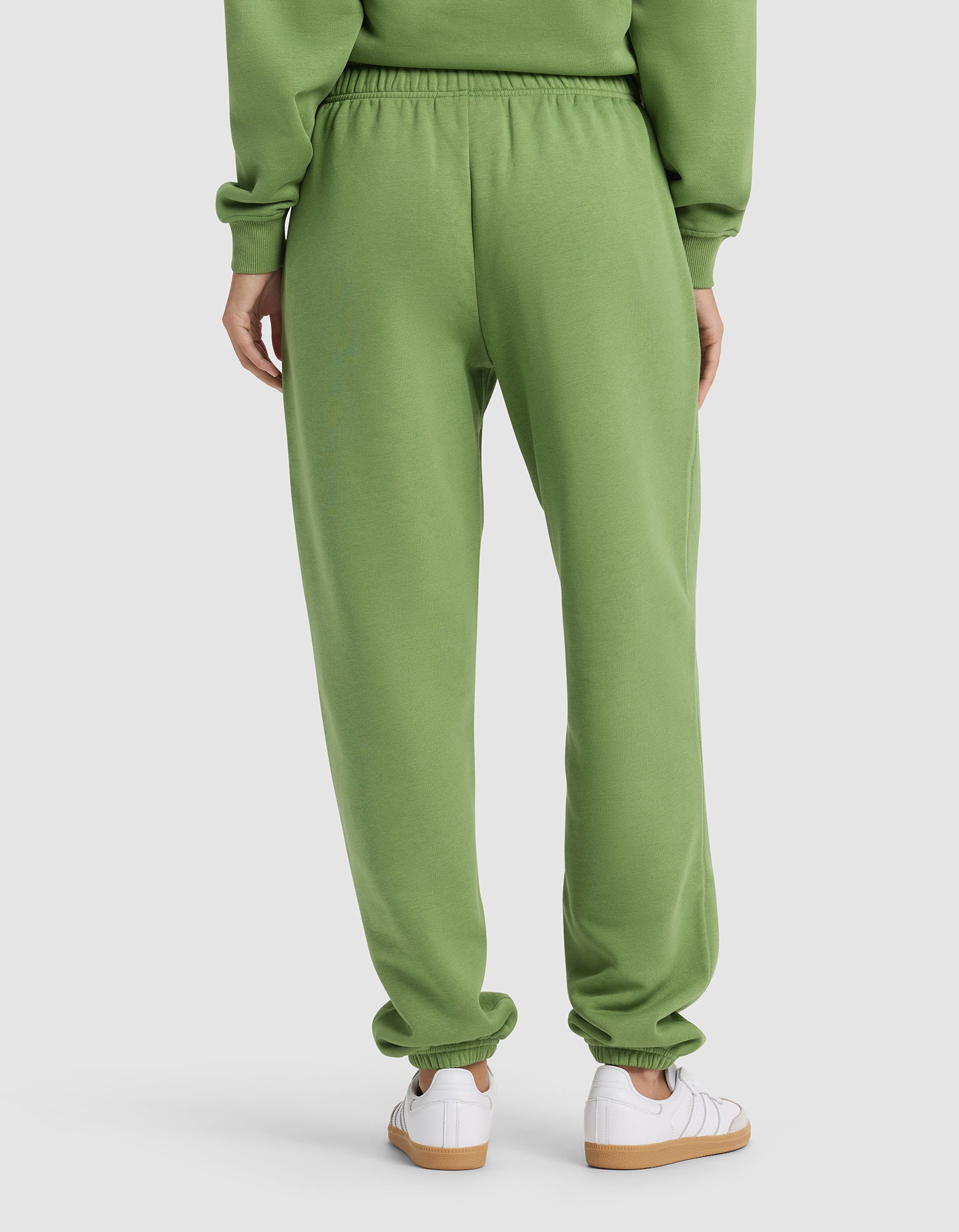 A woman is seen from behind in Cozy Earth's Women's Cityscape 2.0 Sweatpant and a matching green sweatshirt, paired with white sneakers, against a plain background.