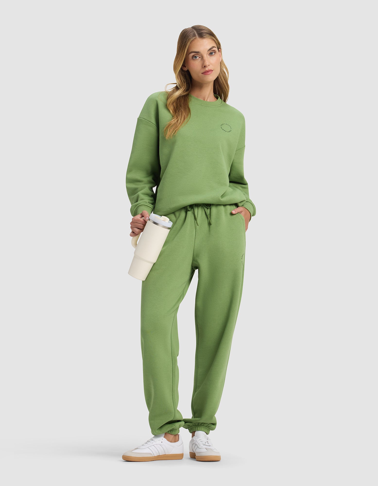 A woman models the Cozy Earth Women's Cityscape 2.0 Sweatpant in green with a matching sweatshirt, white sneakers, and a cream water bottle, posing with one hand in her pocket against a plain light background.