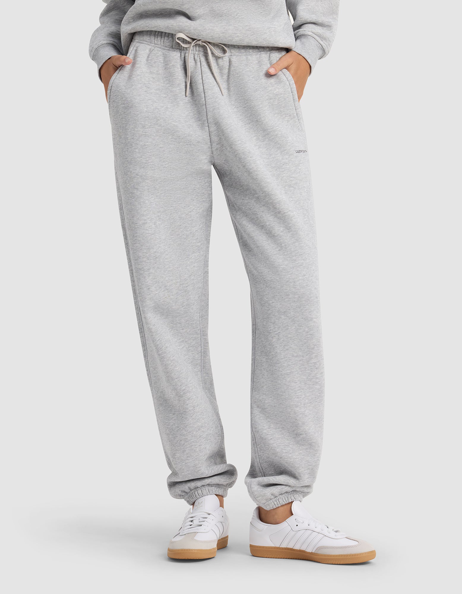 A person wears Cozy Earth's Women's Cityscape 2.0 Sweatpant in light gray with an elastic waistband and drawstring, paired with white sneakers and a matching sweatshirt, hands in pockets.|Color:Heather Grey