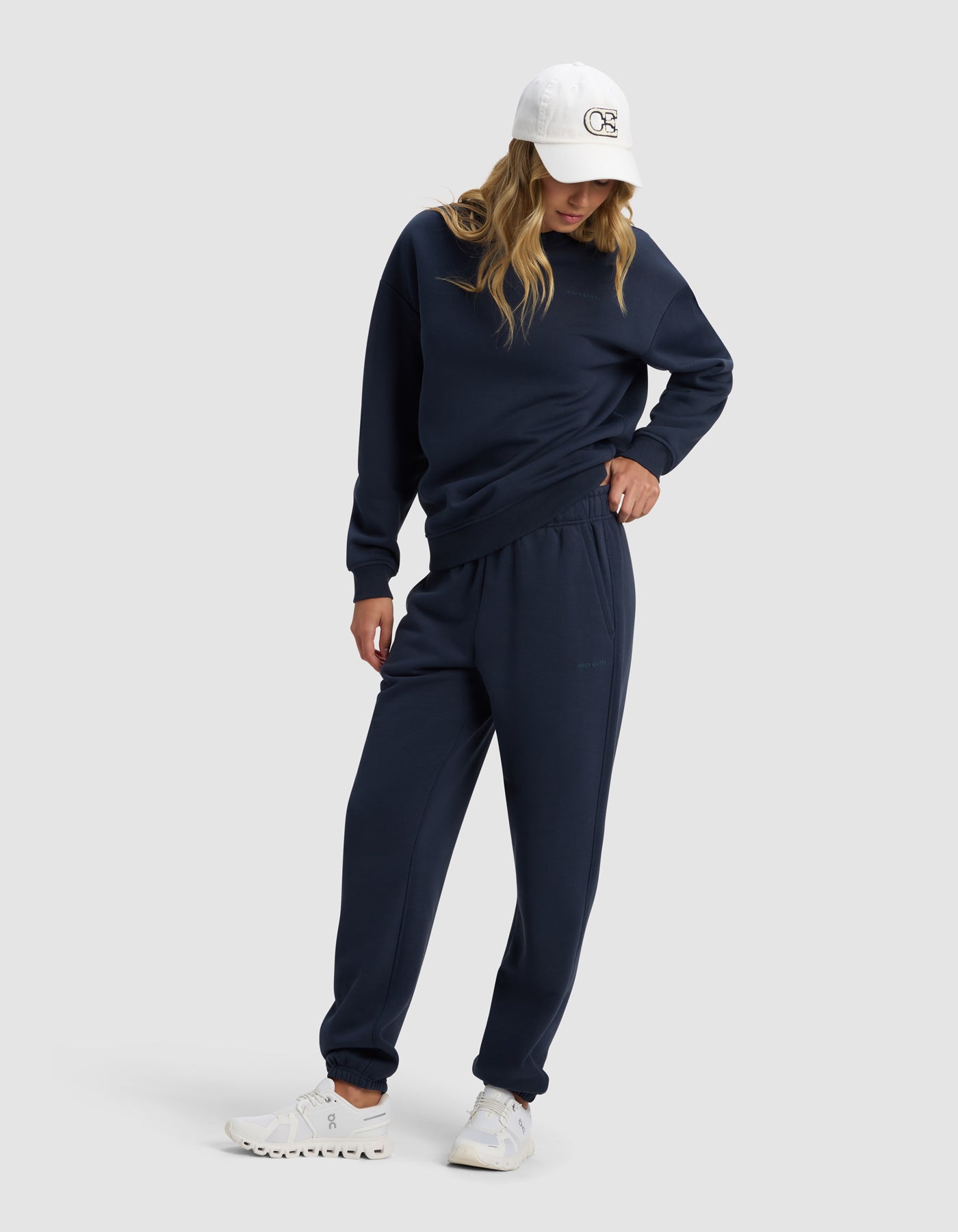 A model in the Cozy Earth Women's Cityscape 2.0 Crewneck and matching sweatpants, styled with white sneakers and a white baseball cap, stands against a light gray background, looking down with one hand in her pocket.