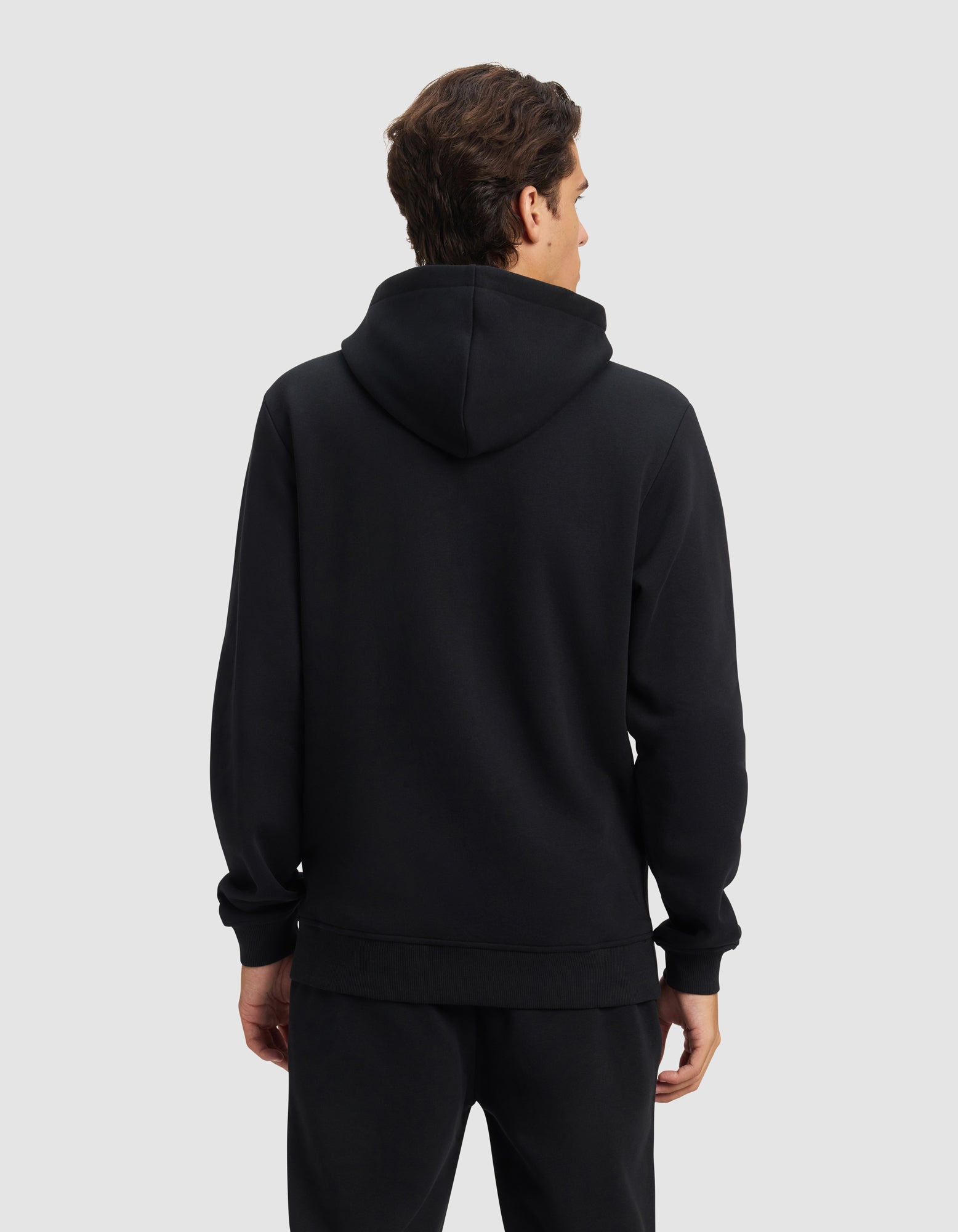 A person with short brown hair faces away, wearing the Cozy Earth Men's Cityscape 2.0 Hoodie and matching black pants against a plain light gray background.