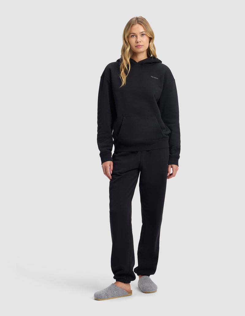 A woman with long blonde hair models the Cozy Earth Women's Cityscape Hoodie & Sweatpant Set in black with grey slippers, standing against a plain light grey background.