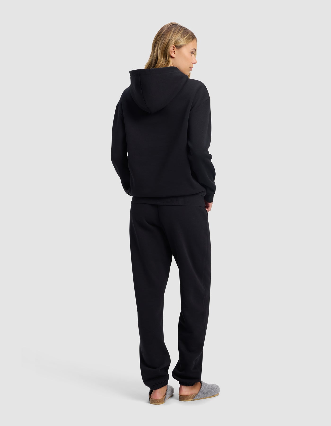 A woman with blonde hair, seen from behind, wears the Cozy Earth Women's Cityscape Hoodie & Sweatpant Set in black with gray slippers, standing against a plain light gray background.|Color:Jet Black