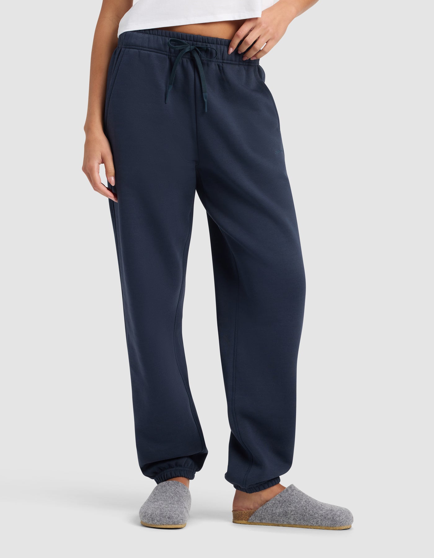 A woman stands against a plain light background wearing Cozy Earth’s Women’s Cityscape 2.0 Sweatpant in navy blue with an elastic waistband and drawstring, paired with a white top and grey slippers.|Color:Eclipse