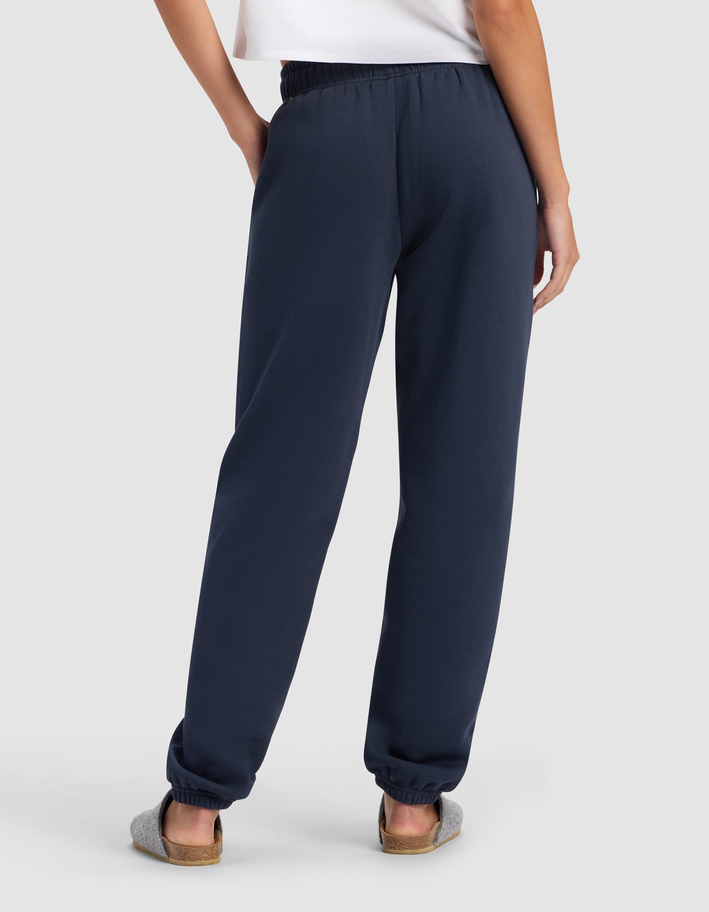 A person stands with their back to the camera, wearing a white shirt and Cozy Earth Women's Cityscape Sweatpant in navy blue, paired with gray slippers against a light background.|Color:Eclipse