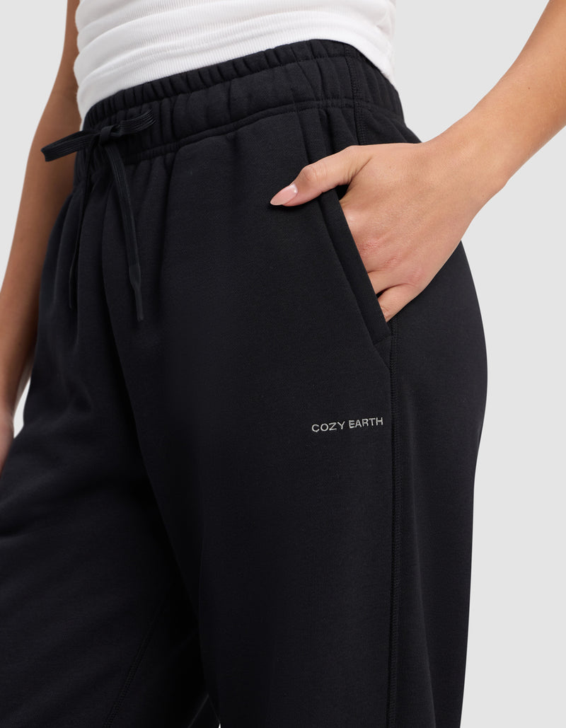 A person wearing black Cozy Earth Women's Cityscape Sweatpants with an elastic waistband, drawstring, a white top, and one hand in the pocket. "COZY EARTH" is printed on the upper thigh of the pants.