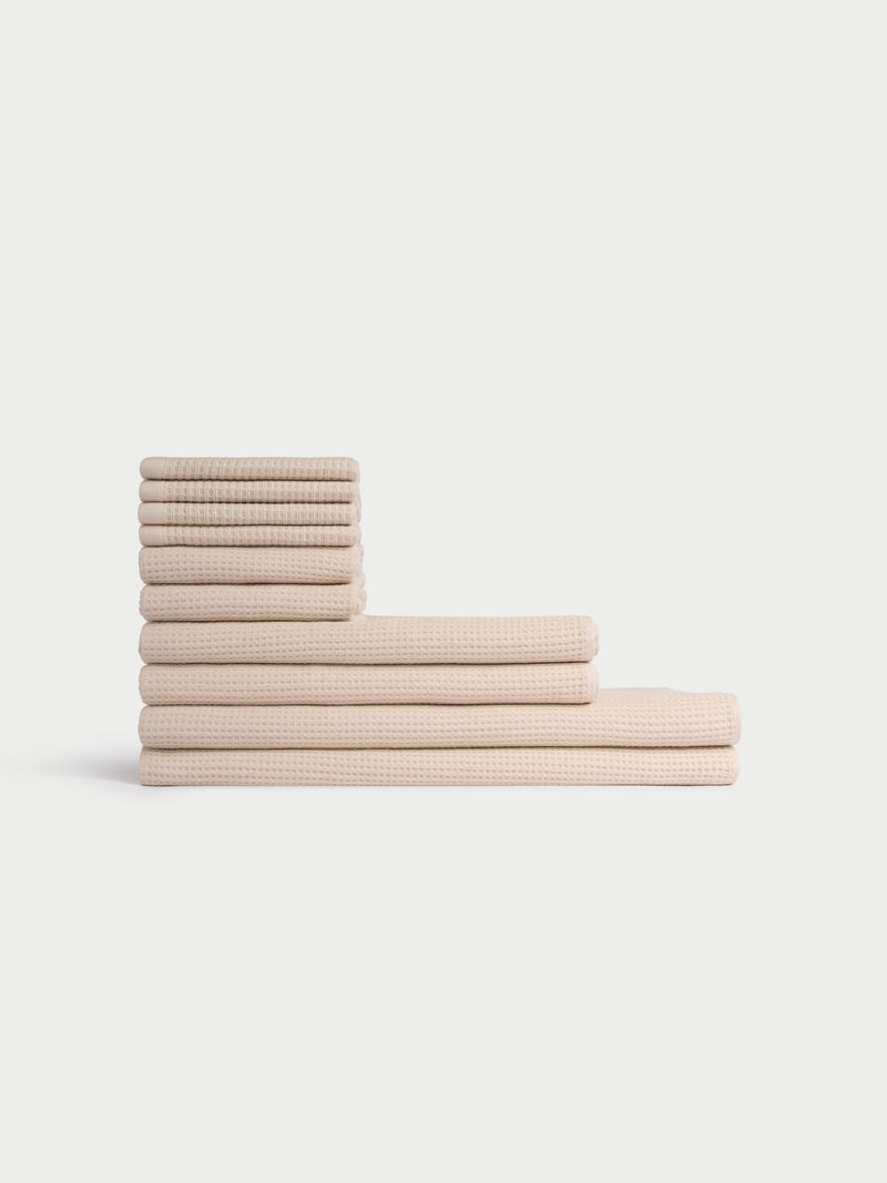 The Cozy Earth Complete Waffle Bath Bundle features beige, waffle-textured towels neatly stacked in two rows on a white background, with smaller towels stacked vertically and larger ones laid horizontally at the bottom.
