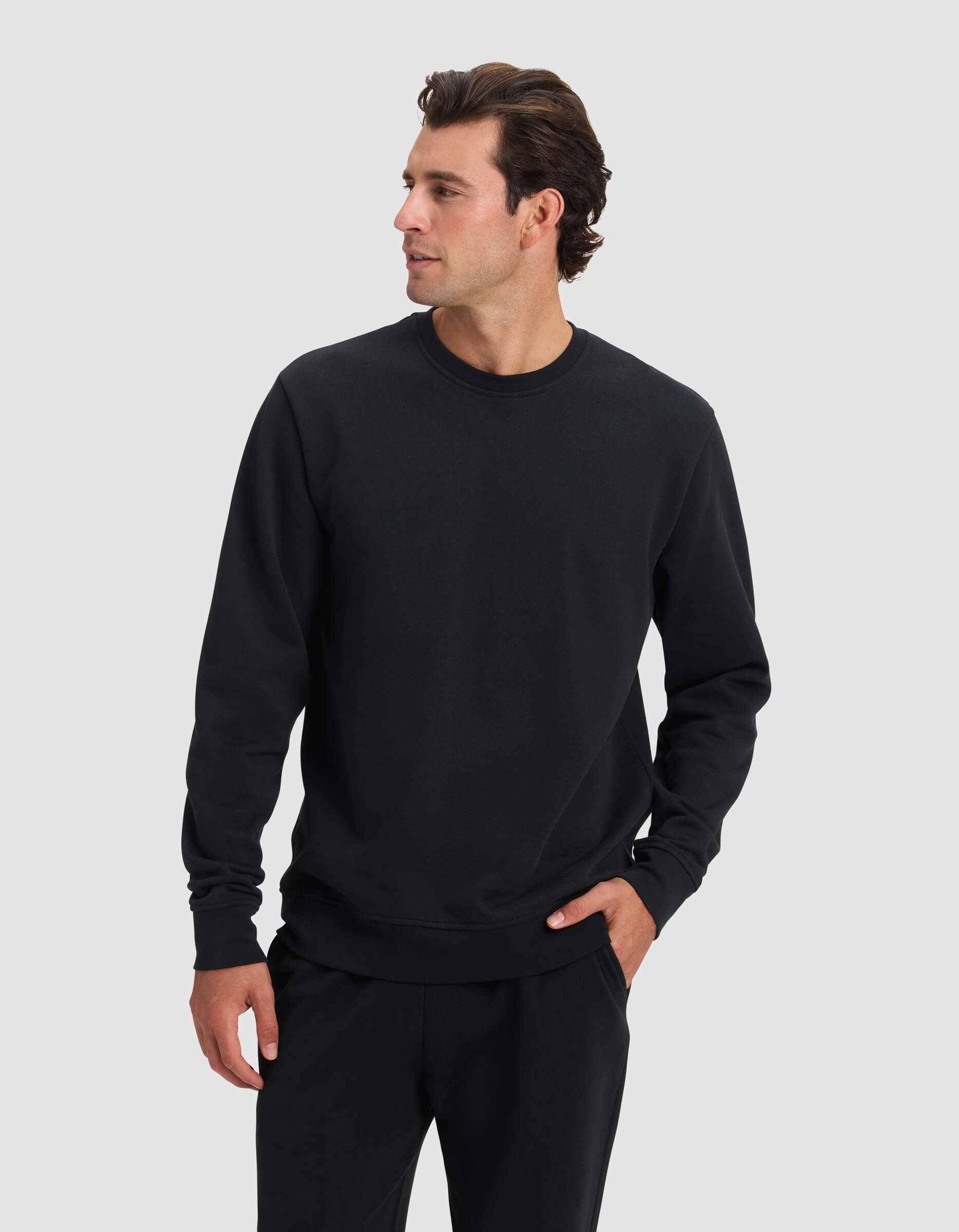 A man wearing the Cozy Earth Men's Dylan Crewneck in black and matching black pants stands against a light gray background, looking to his left with one hand in his pocket.|Color:Jet Black