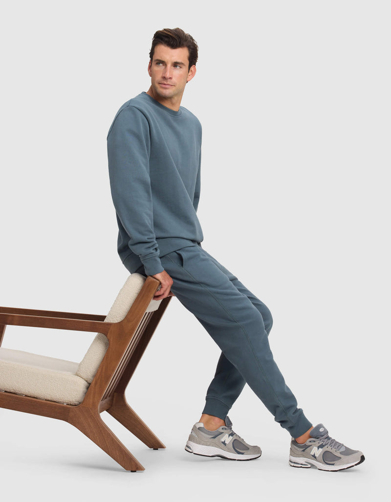A man wears Cozy Earth's Men's Dylan Jogger in teal with a matching sweatshirt, paired with gray New Balance sneakers, as he sits on the backrest of a wooden chair with cream cushions, looking left against a plain white background.