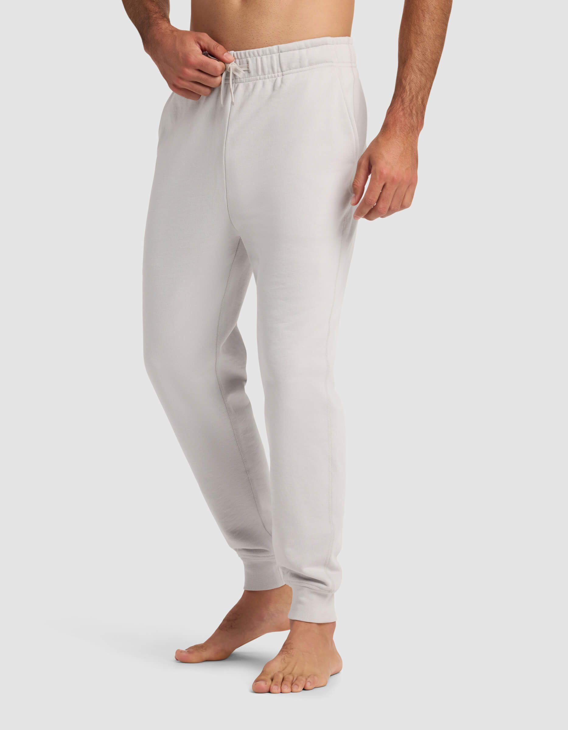 A barefoot man wears Cozy Earth's Men's Dylan Jogger in light gray, featuring an elastic waistband and drawstring, with one hand in his pocket. The background is plain white.