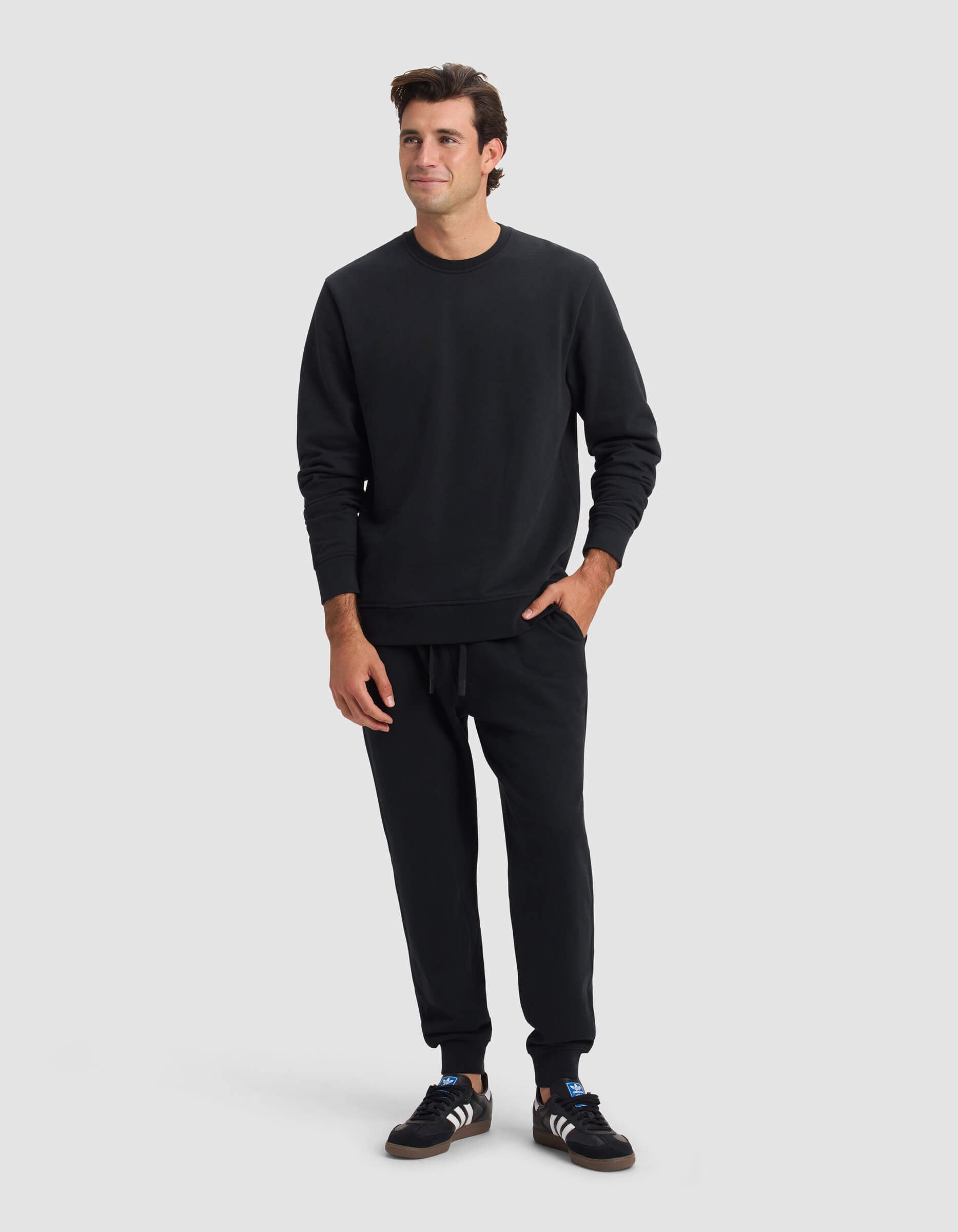 A man stands against a plain light gray background, wearing Cozy Earth's Men's Dylan Jogger in black with a matching sweatshirt and Adidas sneakers. He has short dark hair, a slight smile, and one hand in his pocket.