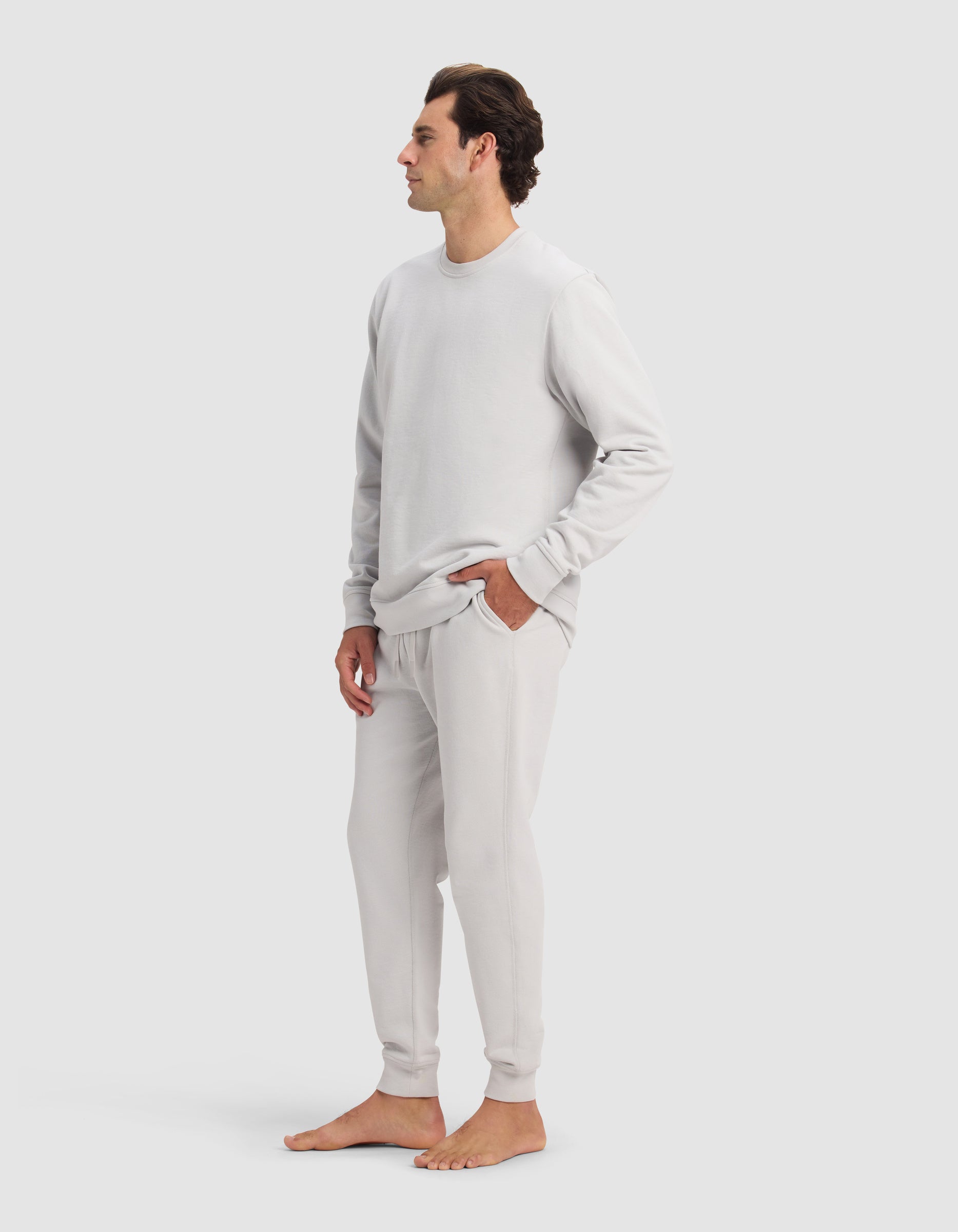 A man stands barefoot with hands in his pockets, wearing the Cozy Earth Men's Dylan Crewneck & Jogger Set in light gray, against a plain light gray background.