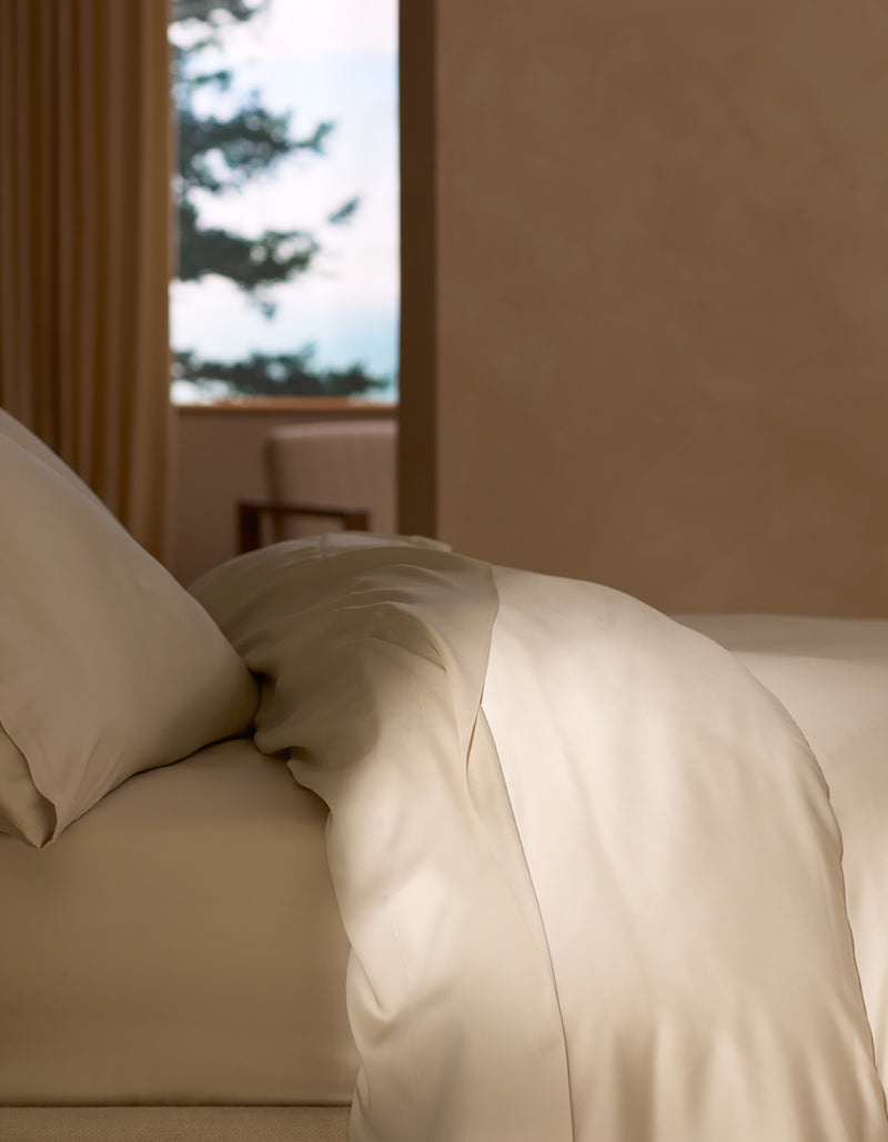 A neatly made bed features the Cozy Earth Bamboo Duvet Cover and matching cream-colored pillowcases, bathed in soft natural light. A window with tree views and a chair near beige walls and curtains complete the serene scene.
